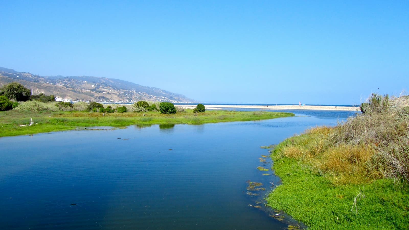 A calm blue lagoon channel between green marsh banks, a sandbar at the ocean in the distance and hills with houses at left