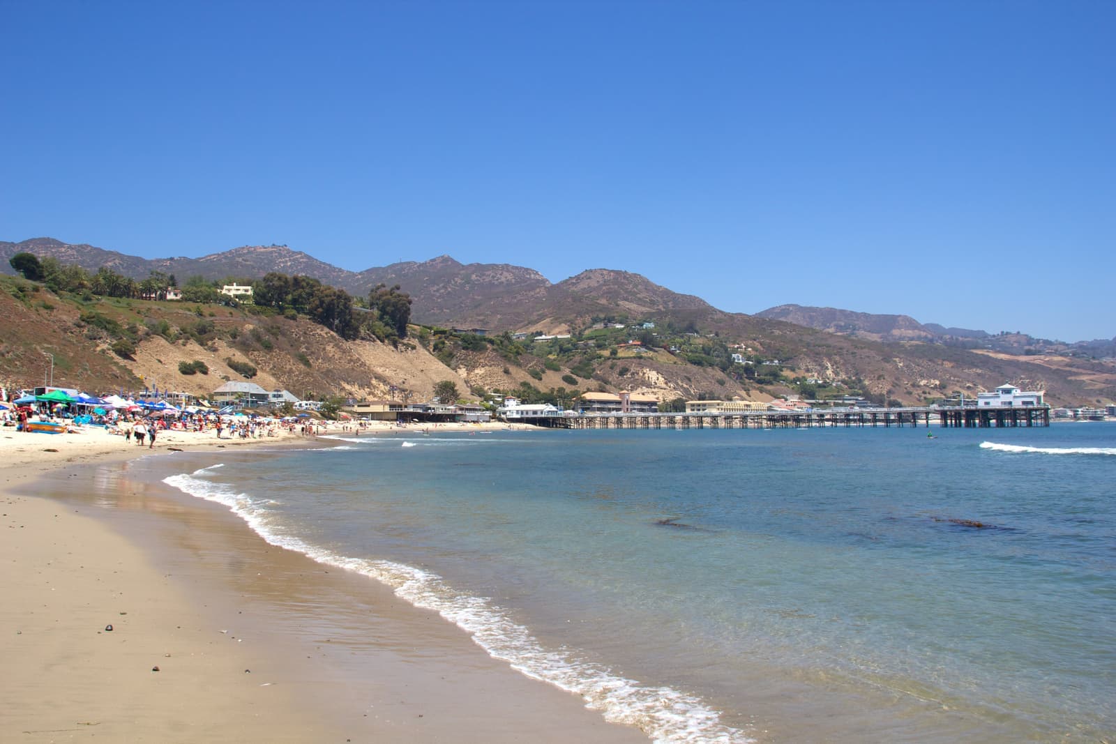 Clear blue water along a curving sandy beach with bright umbrellas at left, a long pier across the bay and dry hills with homes