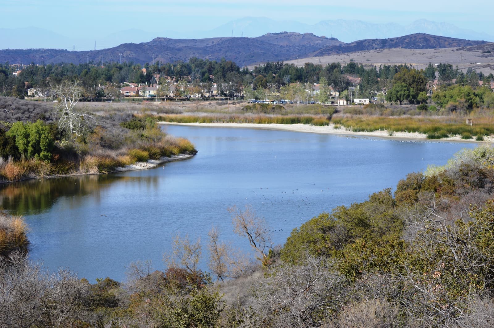 A calm blue reservoir curves between willows and scrub, with tile-roofed homes, trees and brown mountains beyond