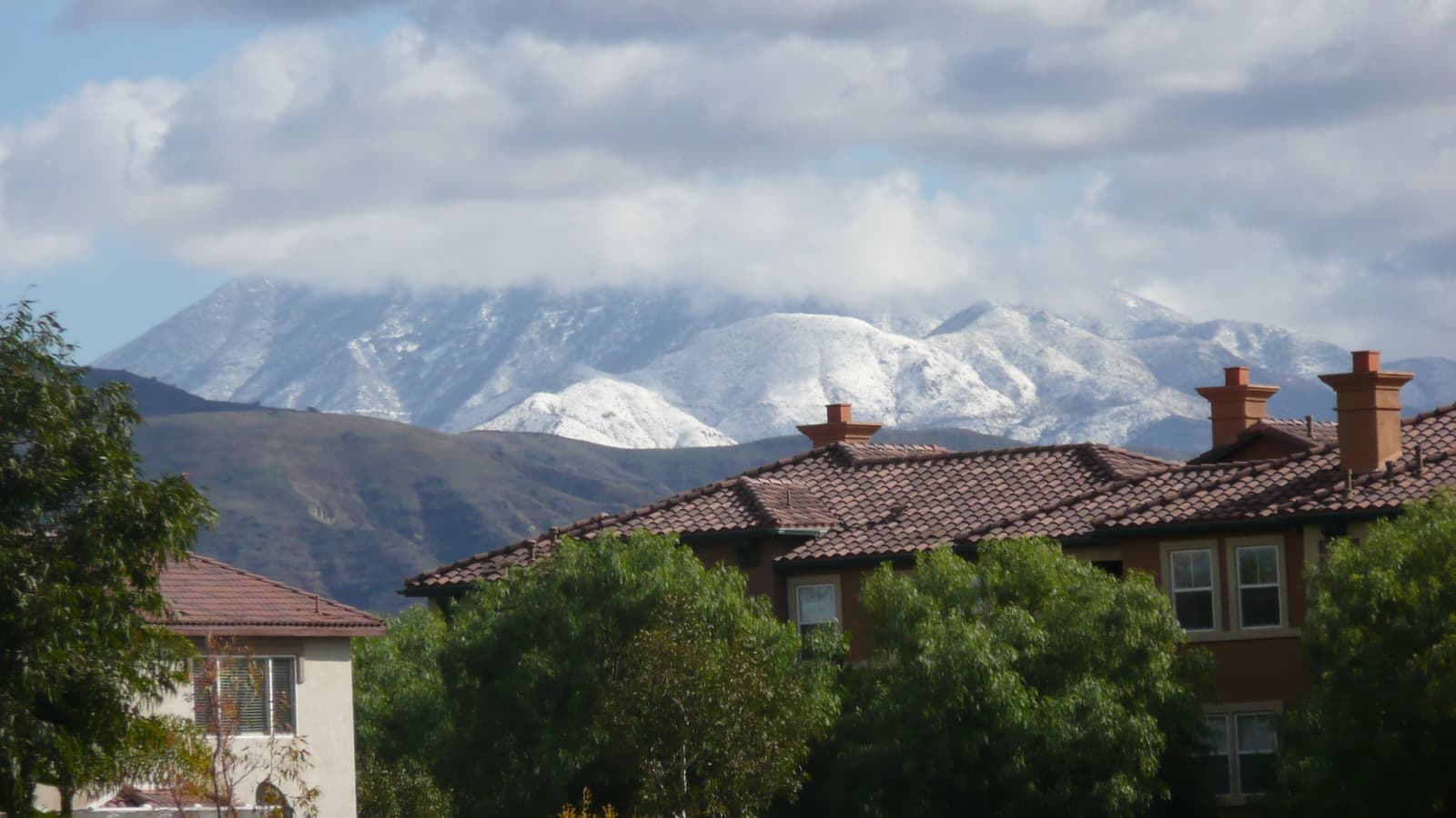 Snow-covered mountain peaks under white clouds rise above green foothills, tile-roofed homes and leafy trees