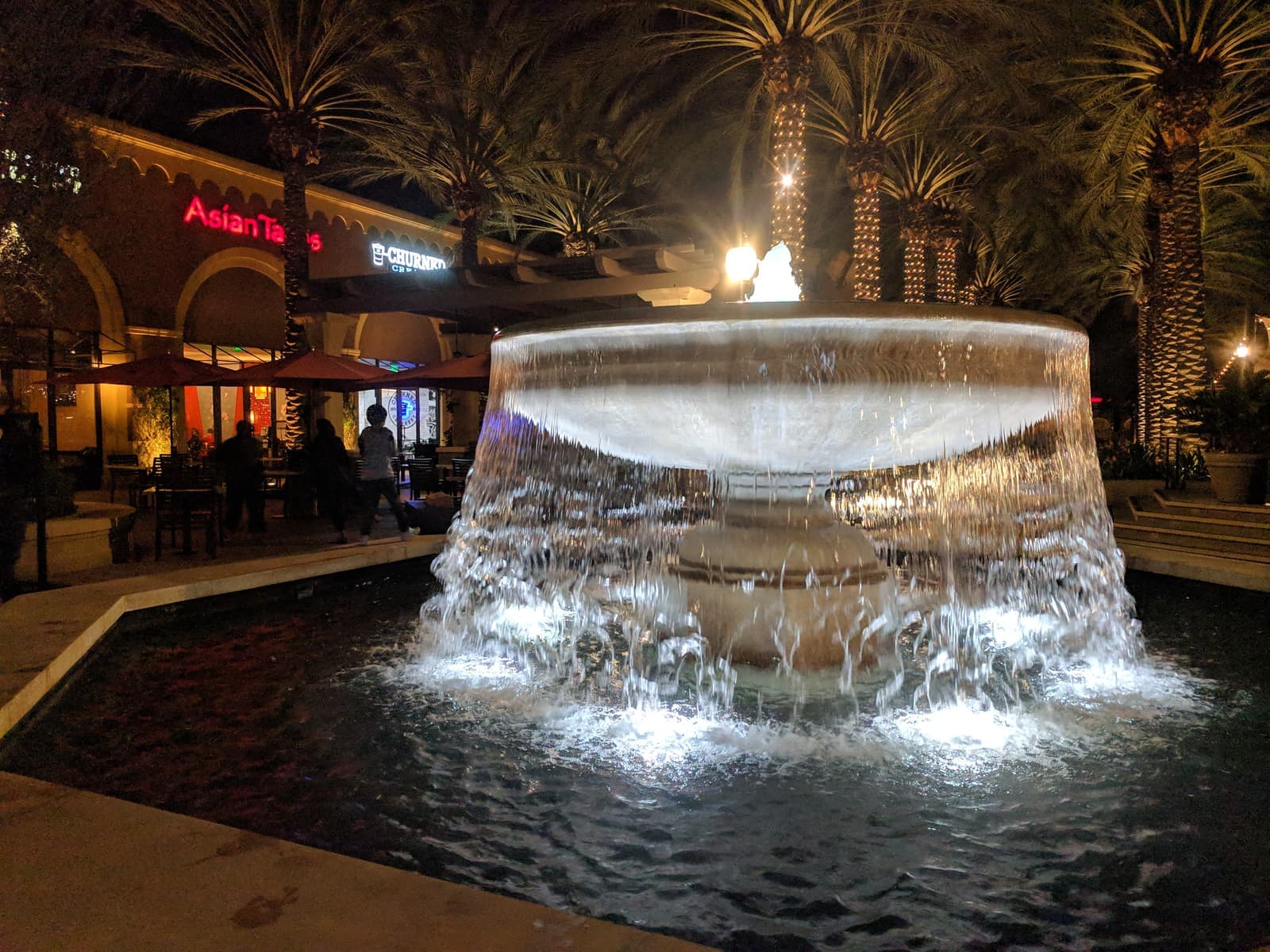 A lit fountain spills water into a pool at night, with palm trees wrapped in string lights and restaurant patios behind
