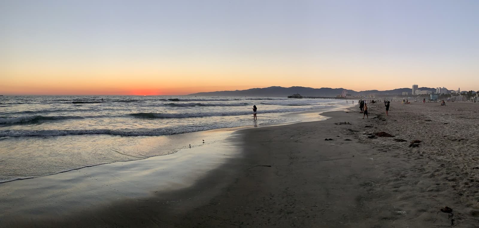 The beach at dusk with small waves, and the Santa Monica Pier and mountains in the distance