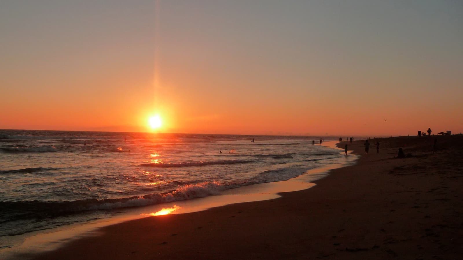 Orange sun low over the ocean at sunset, light reflecting on wet sand, small silhouettes of people along the shoreline