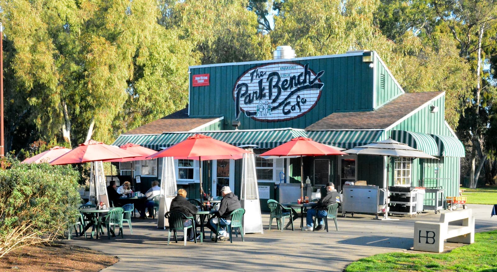 Green wooden cafe with a painted sign and red umbrellas over patio tables, a few people eating outside in low sun among tall trees