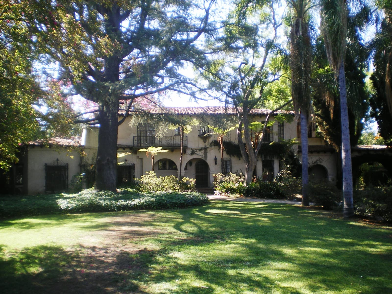 Two story Spanish Revival building with a tile roof, arched doorways and iron balconies behind large trees and a lawn