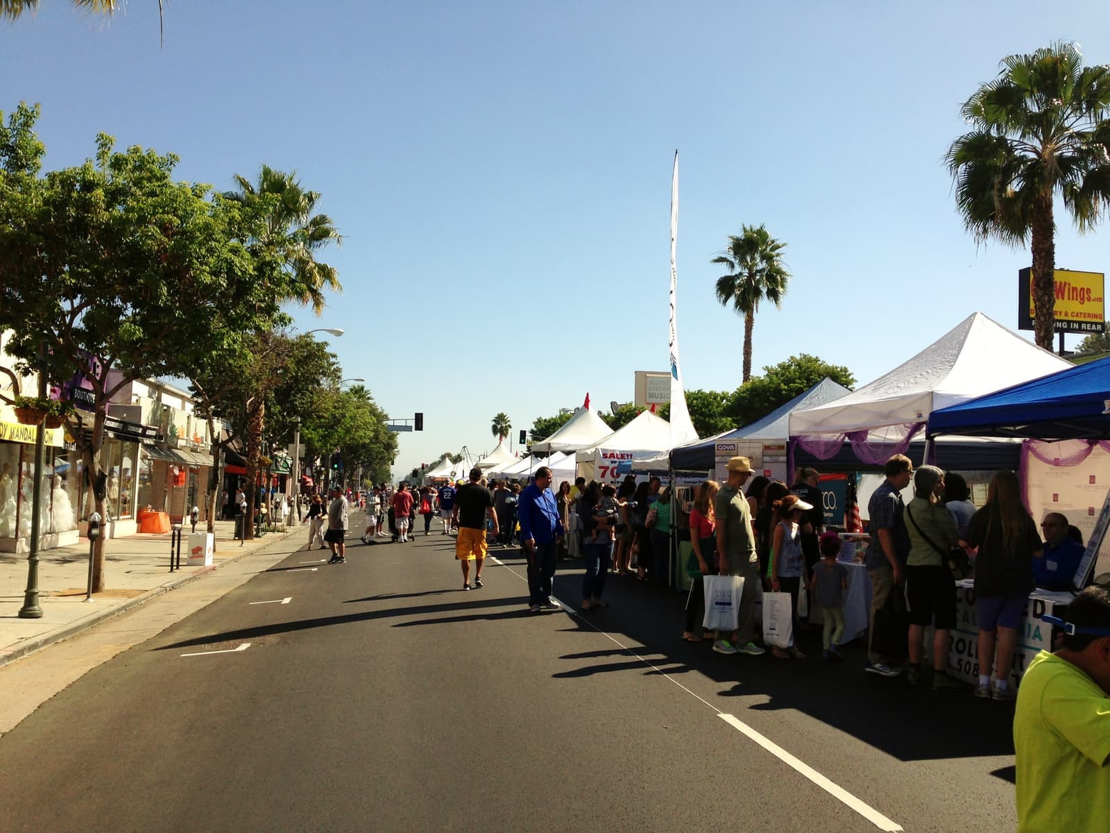 Wide boulevard closed to cars with white vendor tents on one side, people walking in the sun, palm trees and storefronts