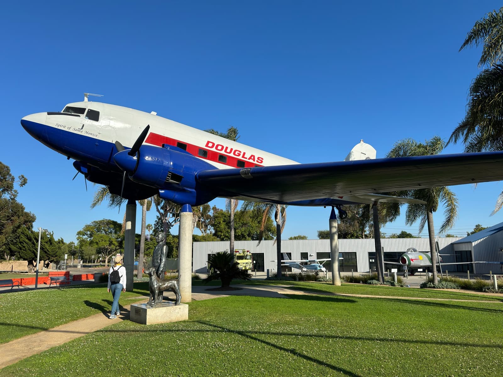Бело-сине-красный Douglas DC-3 на опорах над газоном с пальмами, внизу бронзовая статуя и посетительница, ясное синее небо
