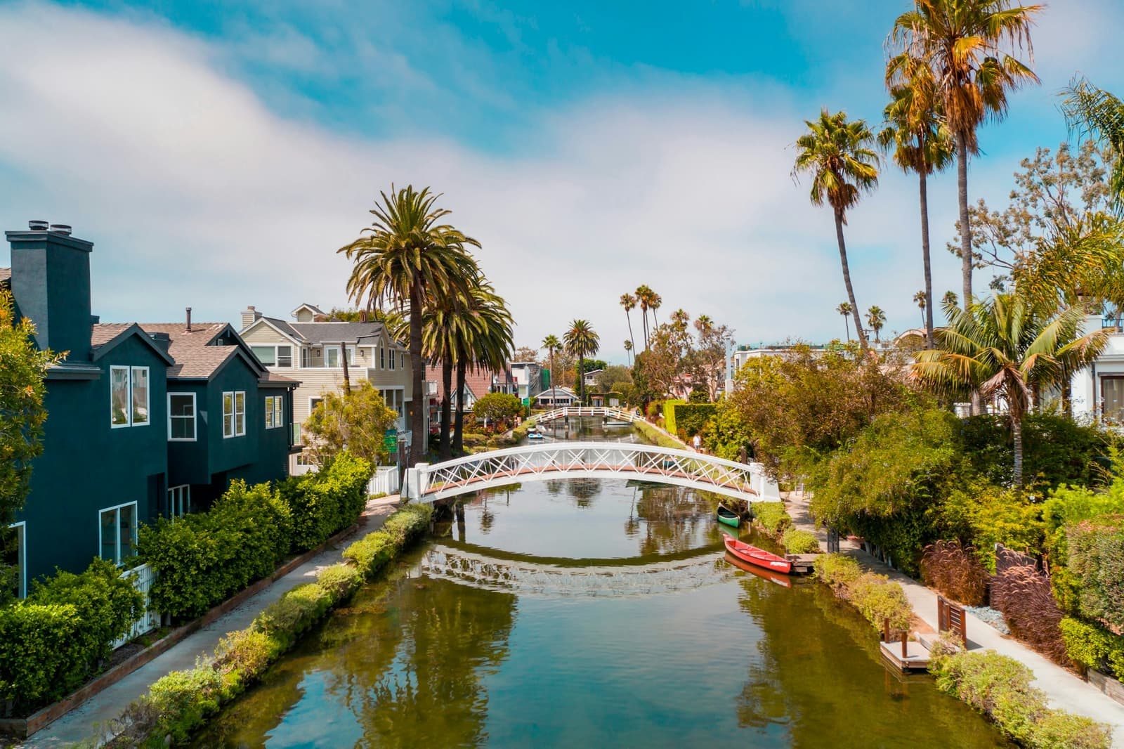 A white arched footbridge over a Venice canal, with palm trees and houses along the water