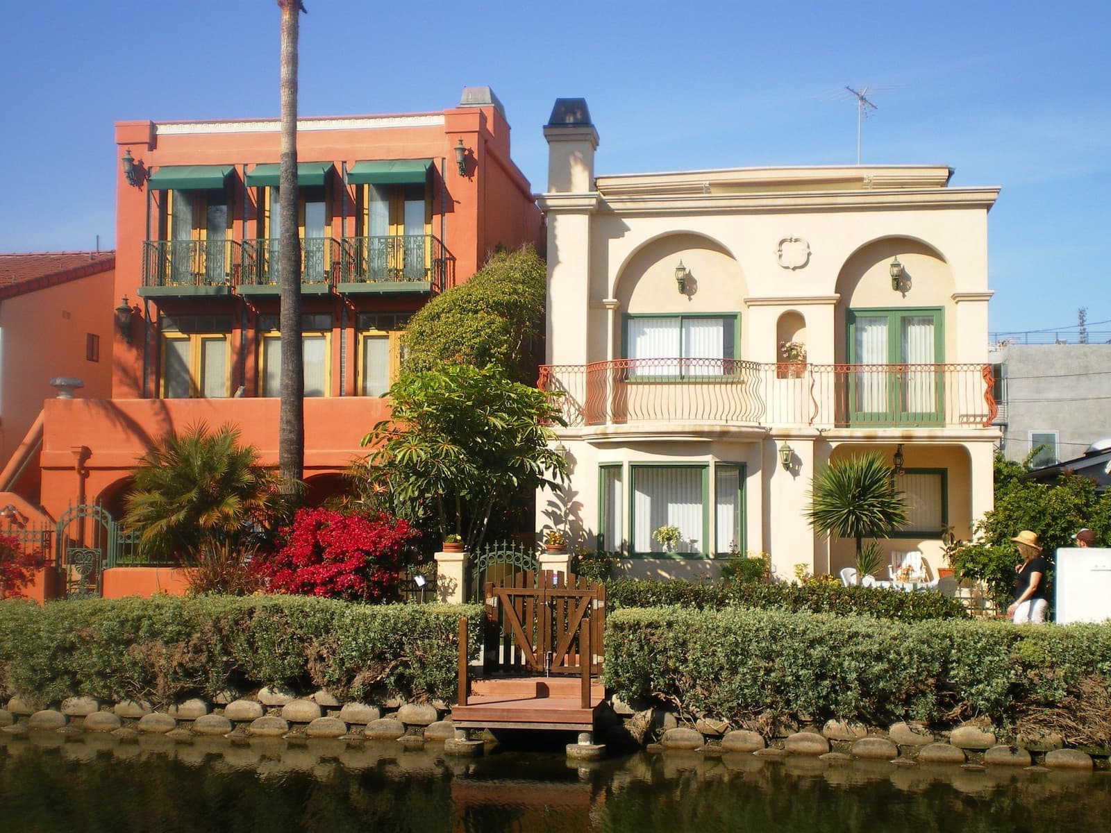A terracotta house and a cream Italianate house with iron balconies behind hedges, red bougainvillea and a small dock on a canal