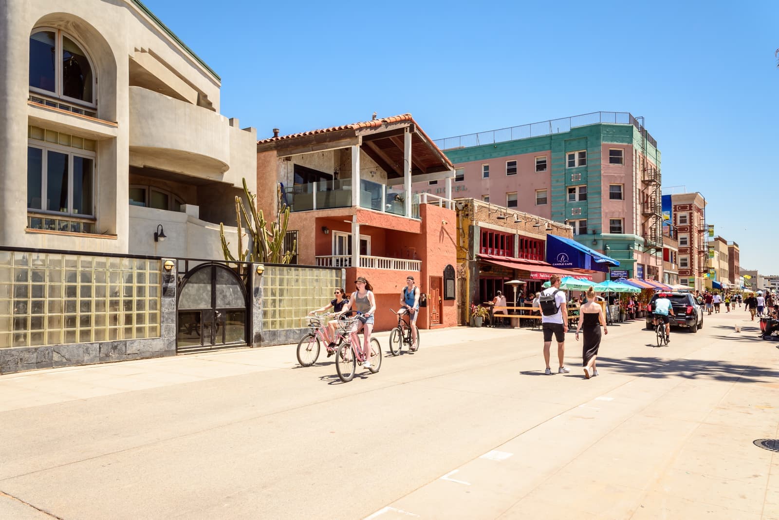People cycling and walking on a wide beachfront walkway past homes, cafe umbrellas and the pink and teal Cadillac Hotel