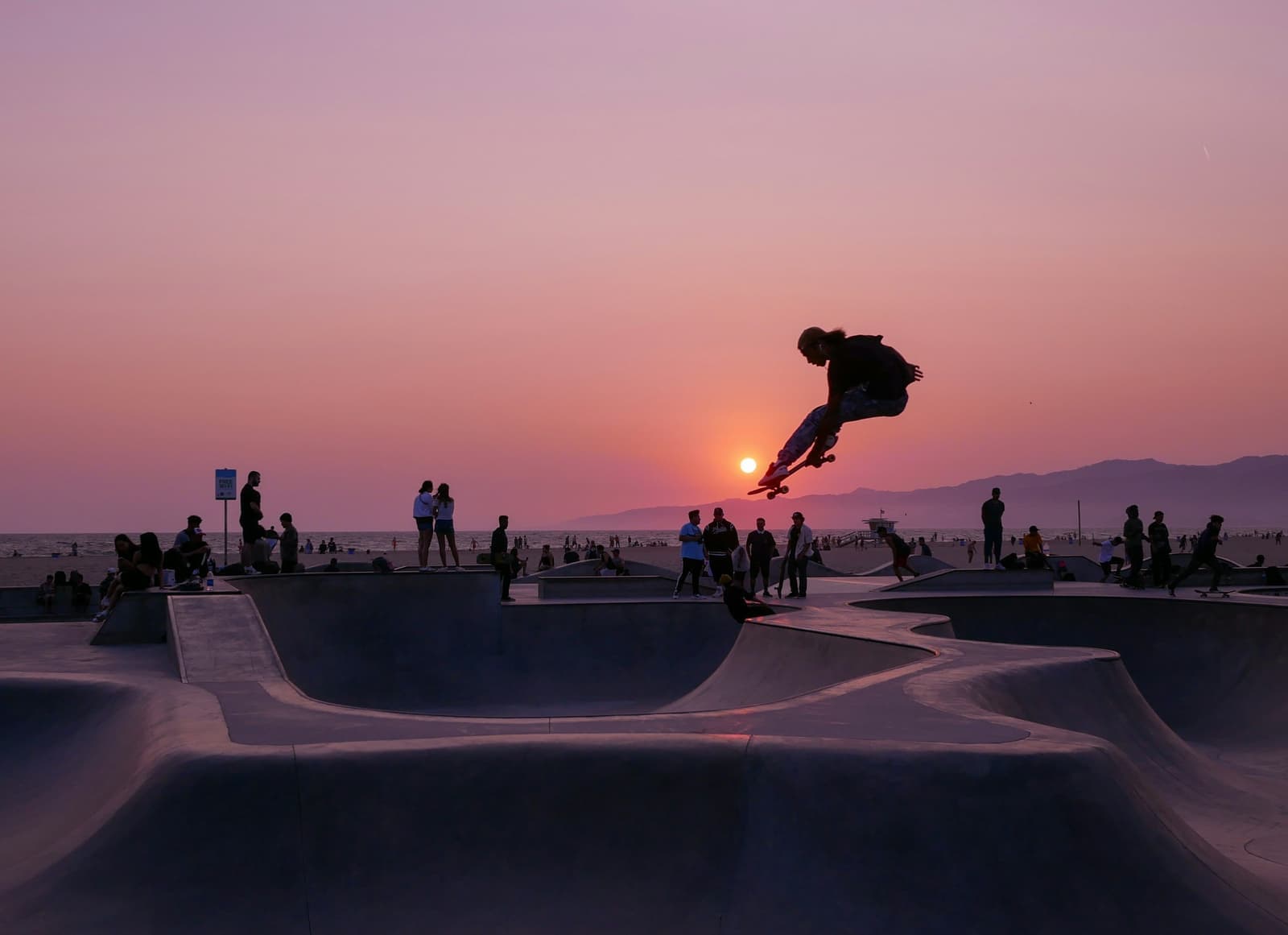 A skateboarder in the air over the Venice Skatepark bowls at sunset, with people watching from the rim