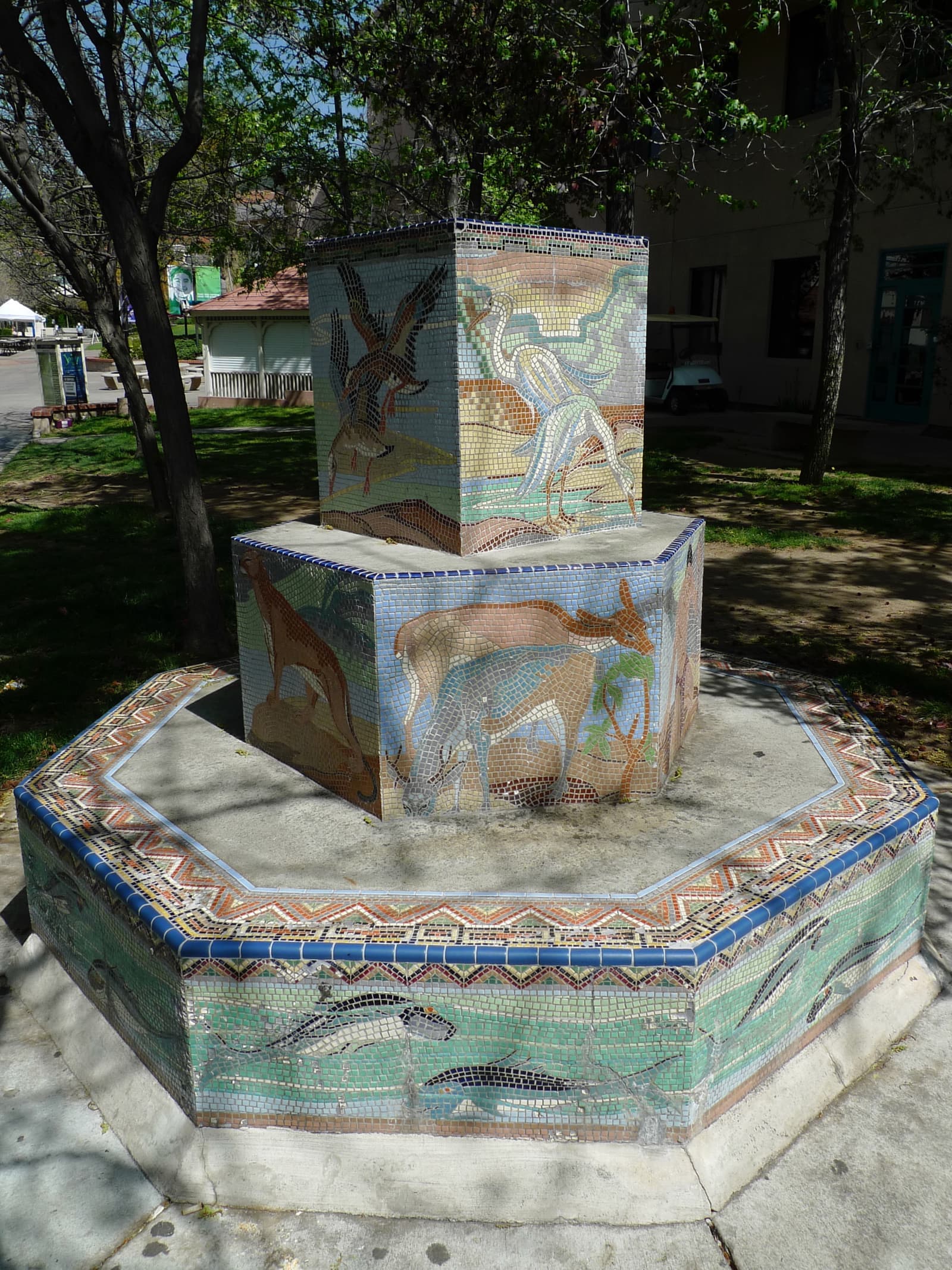 Octagonal fountain covered in colorful mosaic tiles of herons, deer and fish, in dappled shade under campus trees