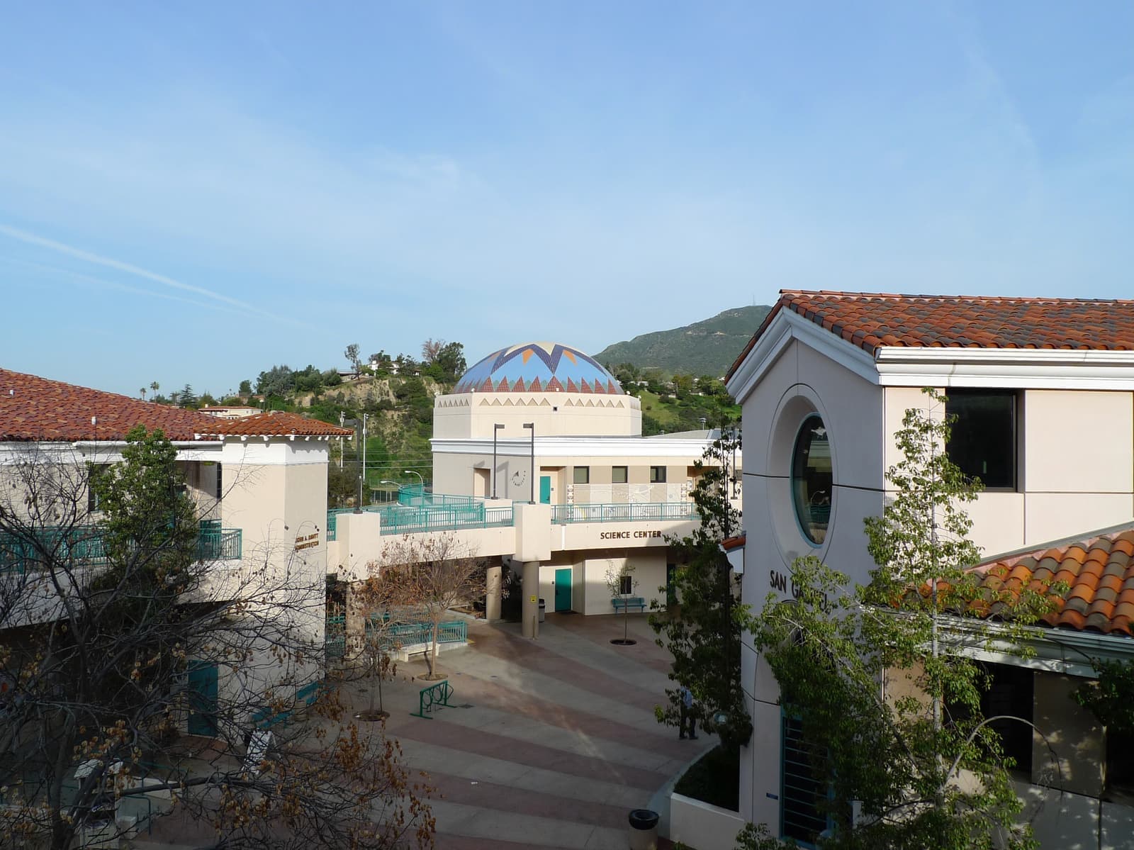 Cream campus buildings with red tile roofs around a paved plaza, a blue and white tiled dome and green hills behind, early morning