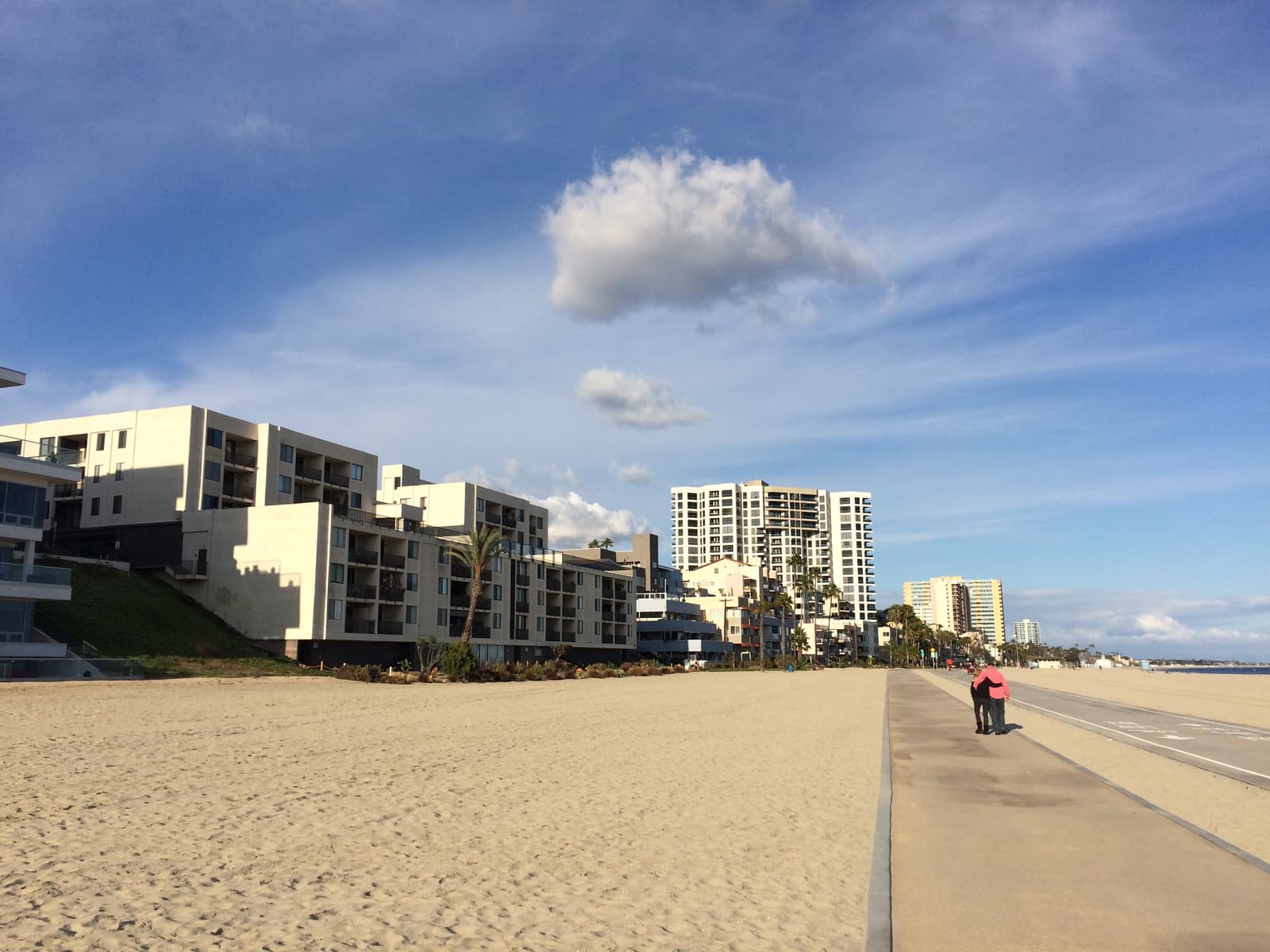 A paved path beside wide sand, lined by low white condo buildings and taller towers under a blue sky, with two people walking