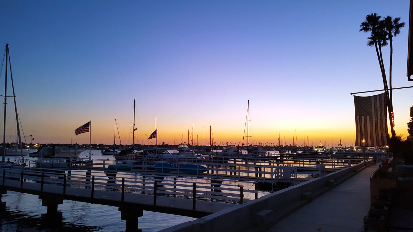 A seawall walkway beside private docks and moored sailboats at dusk, an orange glow on the horizon, flags and a tall palm on the right