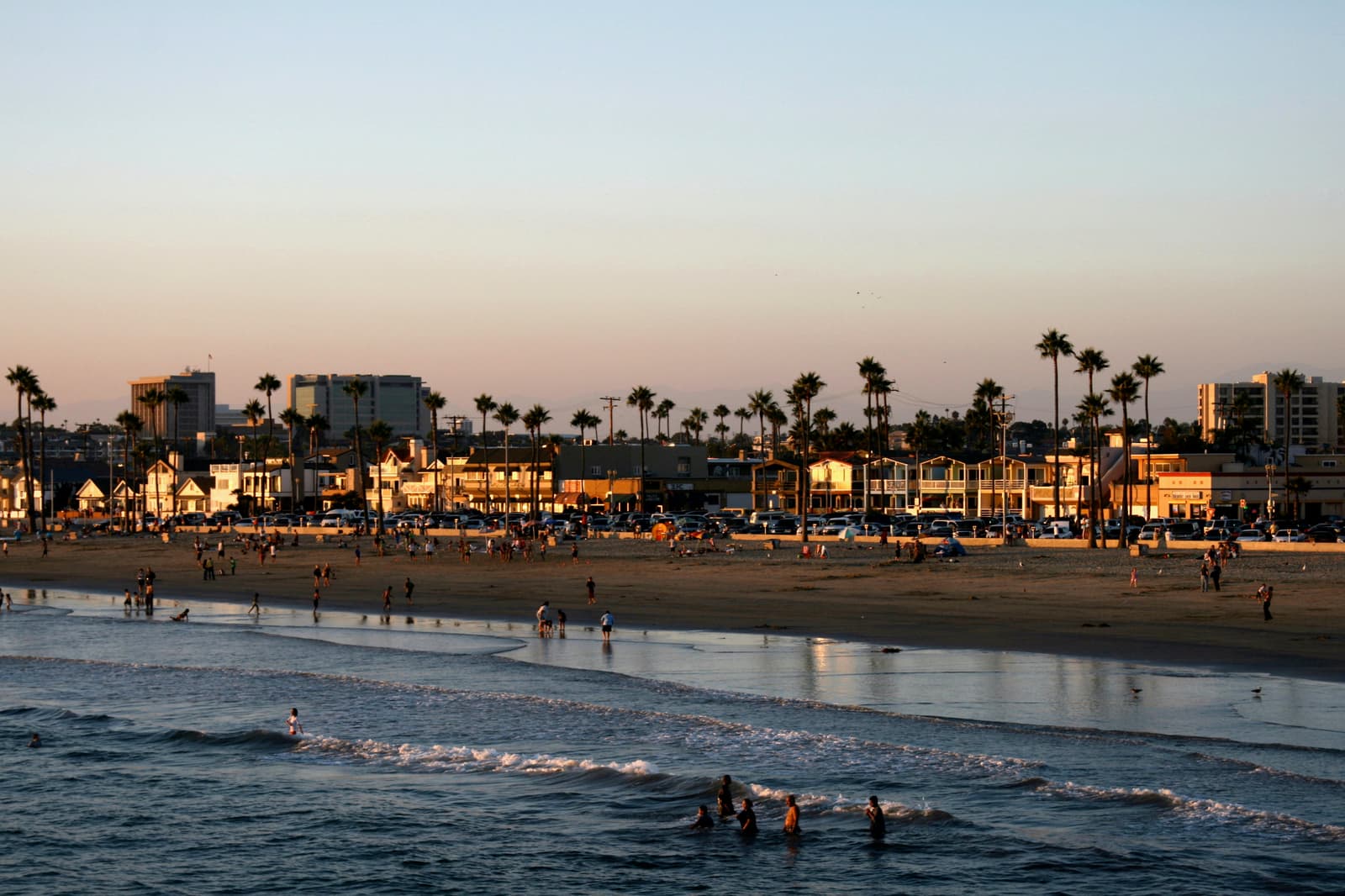 Swimmers in low surf and people on a wide beach in front of a row of beach houses and tall palms lit by warm evening sun