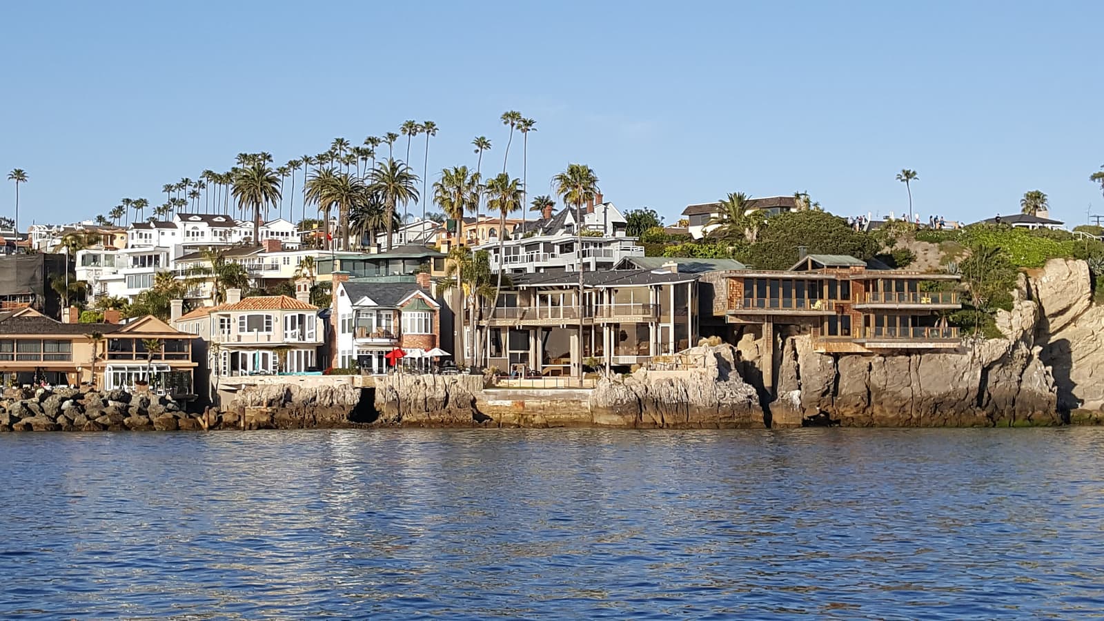 Waterfront houses stacked on a rocky shore under tall palms, seen across calm blue water on a clear day