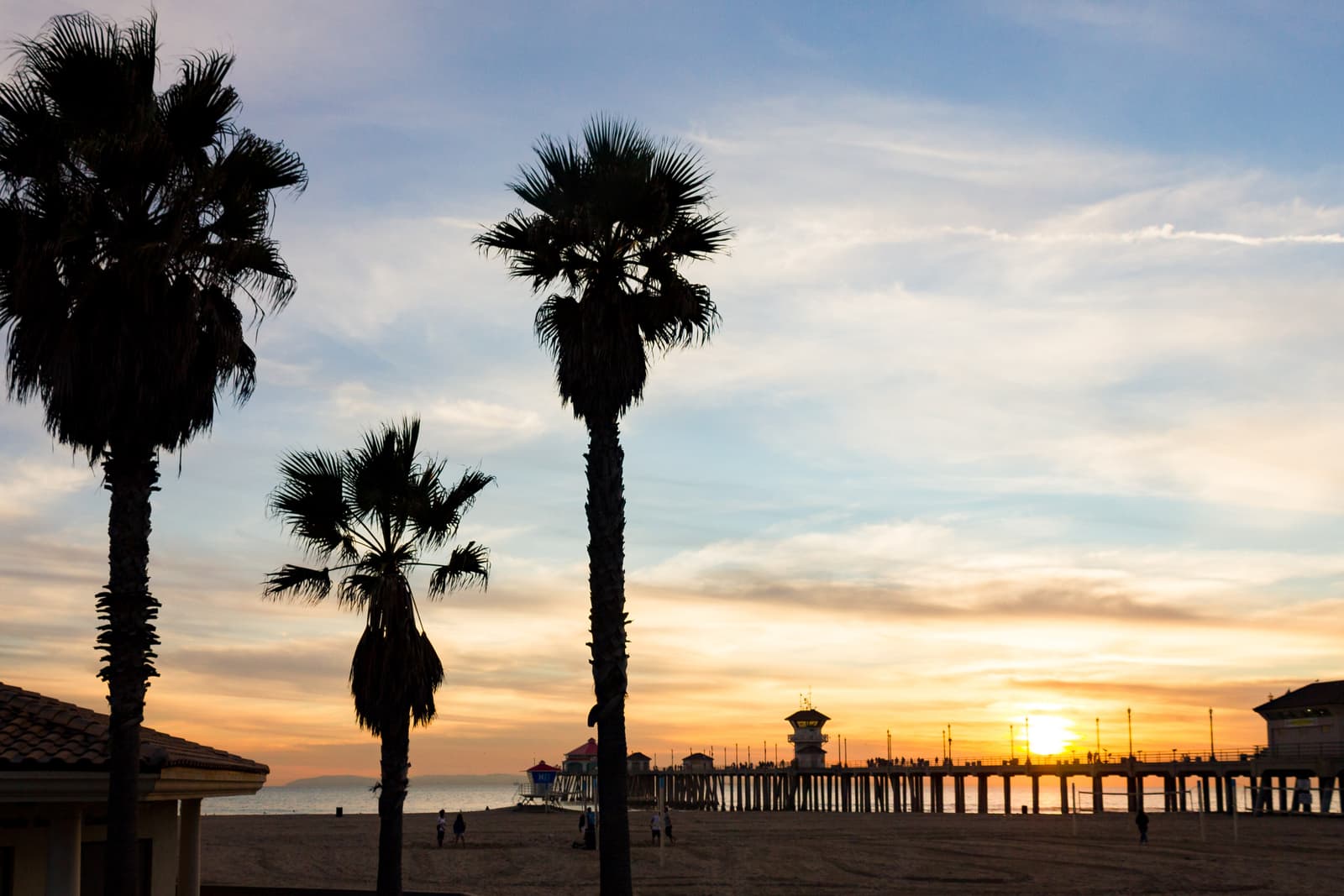 Silhouetted palm trees in front of the long pier and its tower at sunset, the sun low over the water and a few people on the sand
