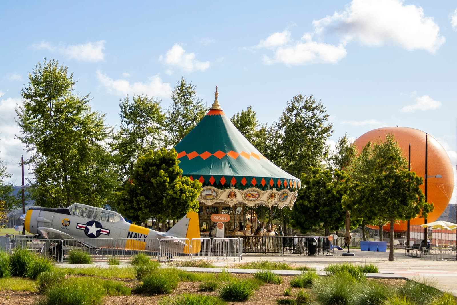 Carousel with a green and orange tent among trees, a silver and yellow vintage Navy plane at left and a big orange balloon at right