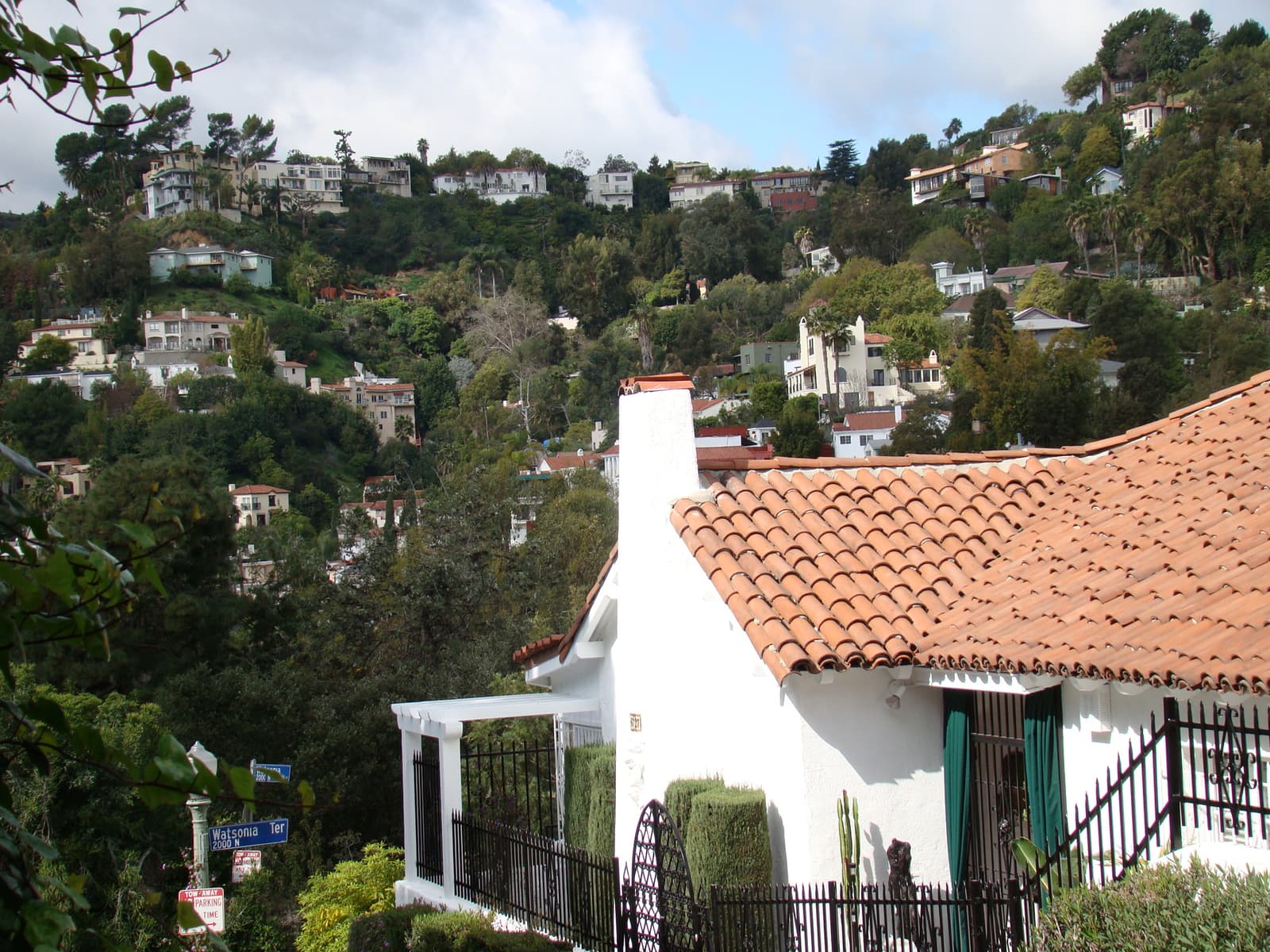 White stucco cottage with a red tile roof and green shutters in front of a steep hillside covered in trees and houses