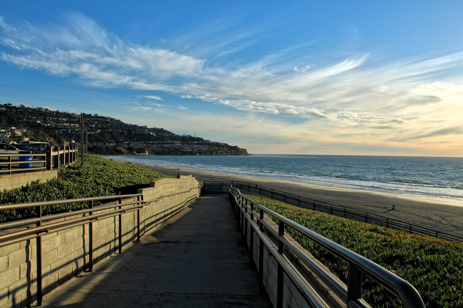 A concrete walkway with metal railings slopes down a green bluff to the beach, with the ocean and Palos Verdes hills under wispy clouds