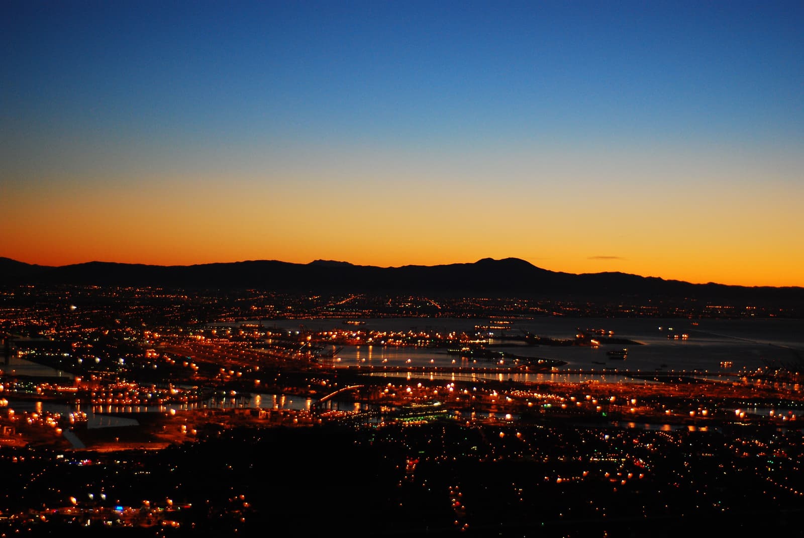 Blue and orange dawn sky over dark mountains, with the lights of the Los Angeles harbor spread out below