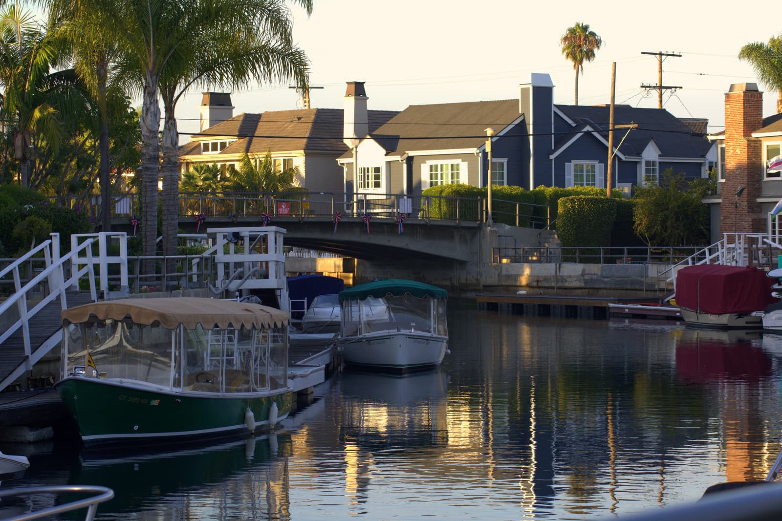 Two canopied electric boats moored on a calm Naples canal at sunset, with homes, palms and a low footbridge behind