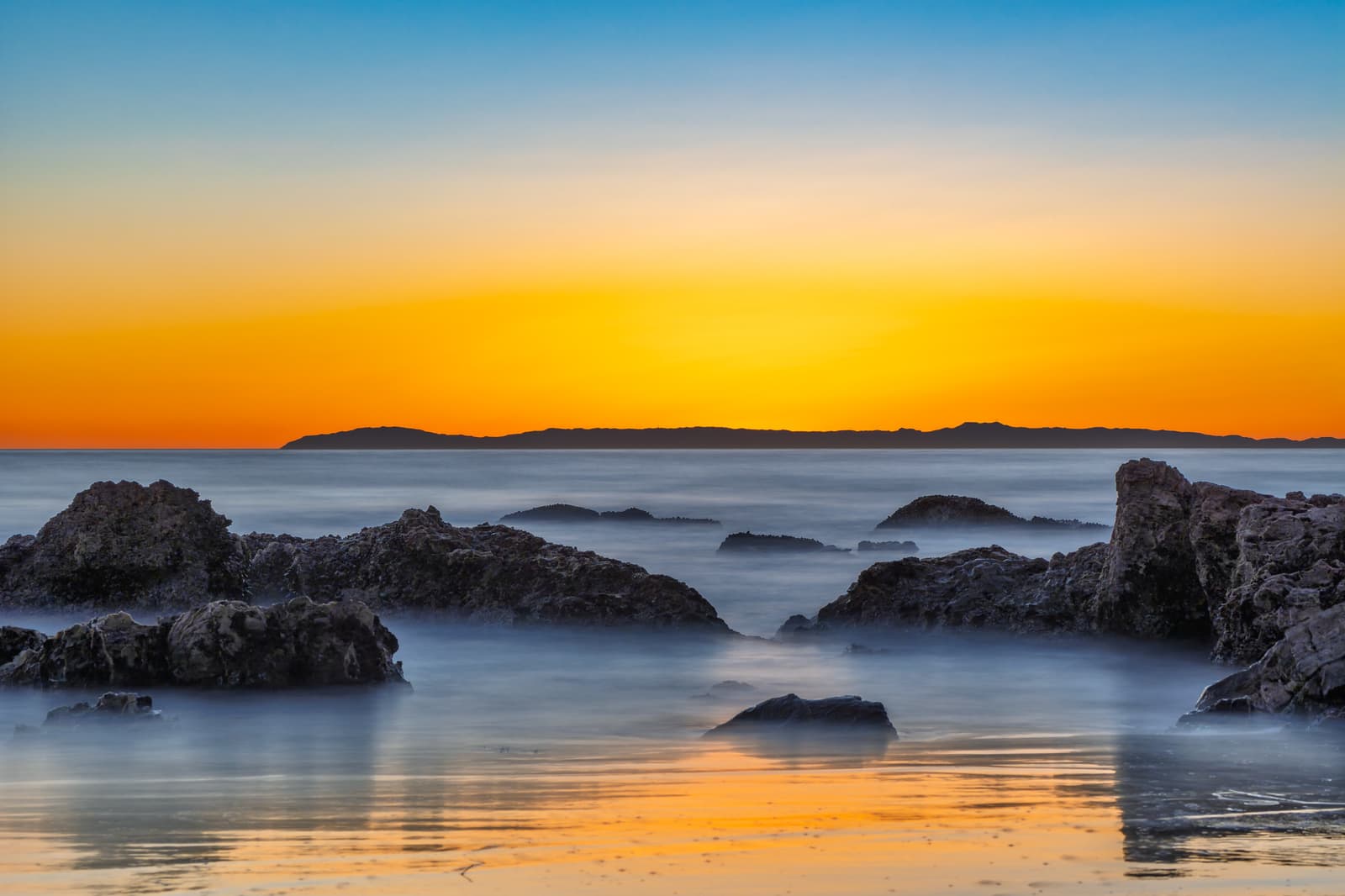 Dark rocks in calm, glassy water under an orange sunset sky, with the long low outline of Catalina Island on the horizon