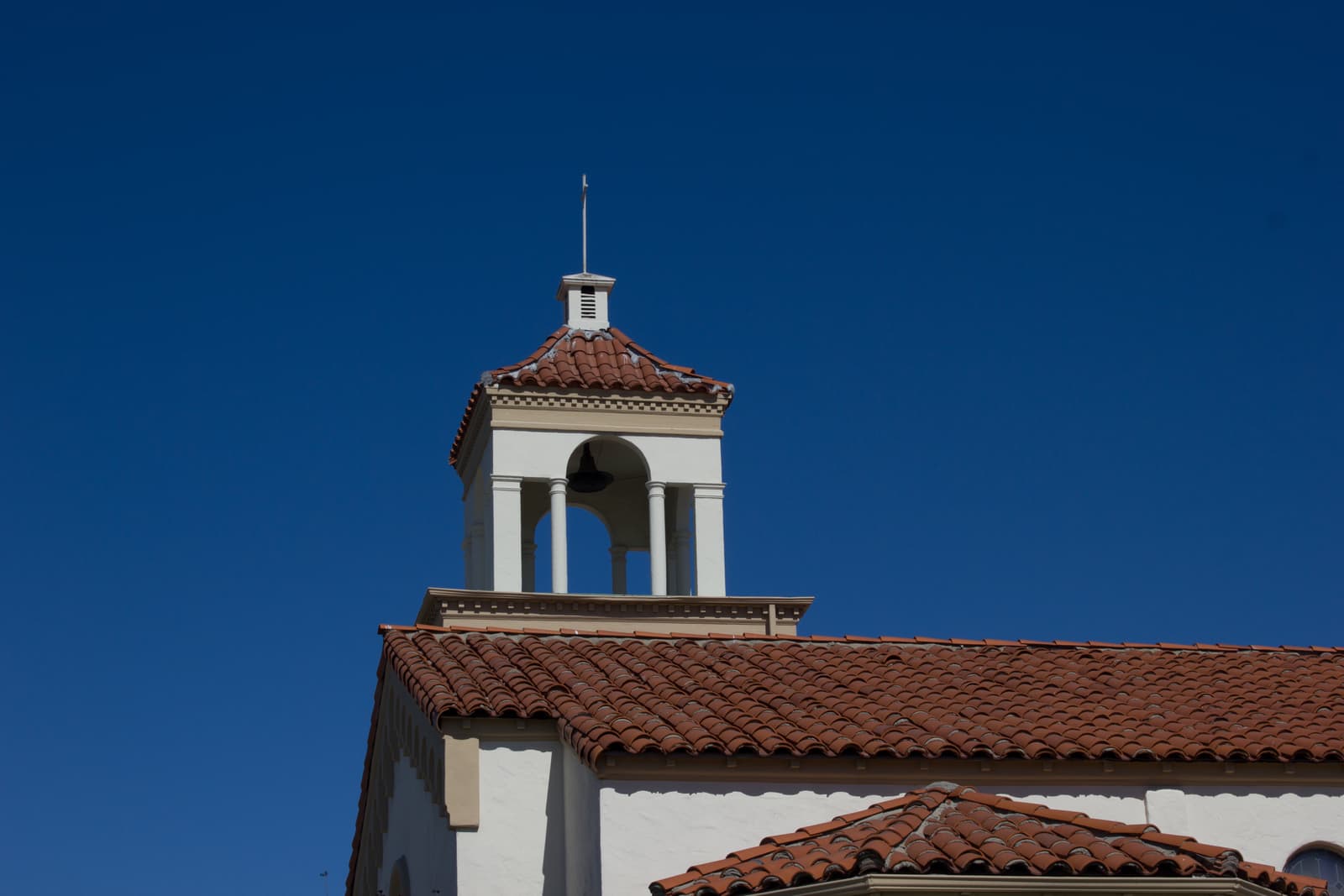 A white bell tower with arched openings and a red tile cap rises above a tiled church roof against a deep blue sky