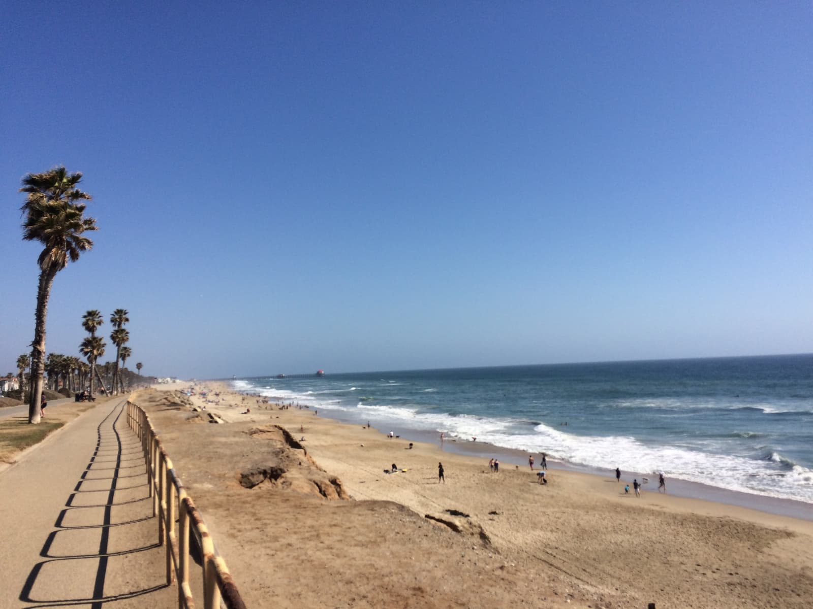 Paved path beside a wooden fence casting long shadows, palm trees ahead and a wide sandy beach with surf and a few people below